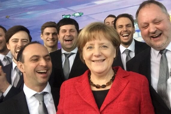 Eine Frau in einer roten Jacke lächelt für ein Selfie mit einer Gruppe von Männern in Anzügen und Krawatten hinter ihr, die alle lachen und lächeln unter einer blau getönten Decke.