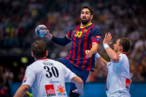 Ein Handballspieler in einem blau-rot gestreiften Trikot mit der Nummer 33 springt zum Schuss, während er den Ball in der Hand hält und zwei Spieler in weißen Trikots versuchen, ihn während eines Spiels zu blockieren.