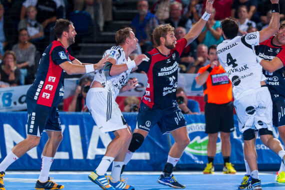 Vier männliche Handballspieler in Aktion auf einem Hallenplatz, zwei in weißen Trikots und zwei in blauen und roten Trikots, mit einer Zuschauermenge im Hintergrund.