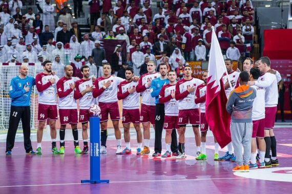 Eine Herren-Handballmannschaft in kastanienbraunen und weißen Trikots steht in einer Reihe auf einem Spielfeld und hält während einer Zeremonie die Hände über dem Herzen. Ein Spieler hält eine Flagge von Katar. Die Zuschauer in Weiß füllen die Tribünen hinter ihnen.
