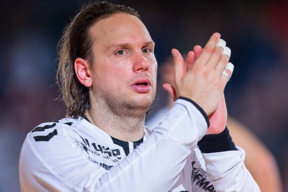 Ein männlicher Handballspieler in einem weißen Trikot mit schwarzen Markierungen klatscht, mit einem konzentrierten Gesichtsausdruck. Er hat schulterlanges Haar und Klebeband an den Fingern. Der Hintergrund ist unscharf.