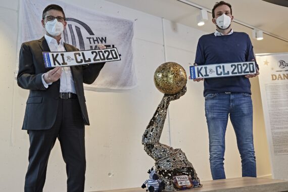Zwei Männer mit Gesichtsmasken halten Nummernschilder mit der Aufschrift KIEL CL 2020 und stehen neben einem Handballpokal. Im Hintergrund sind ein Banner des THW Kiel und eine Danksagungstafel zu sehen.