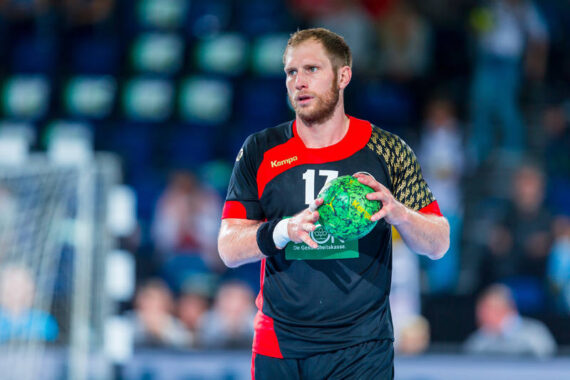 Ein männlicher Handballspieler in einem schwarz-roten Trikot, der einen grünen Handball hält, steht auf einem Hallenplatz und blickt während eines Spiels nach vorne. Im Hintergrund sind unscharfe Zuschauer und ein Teil des Tores zu sehen.