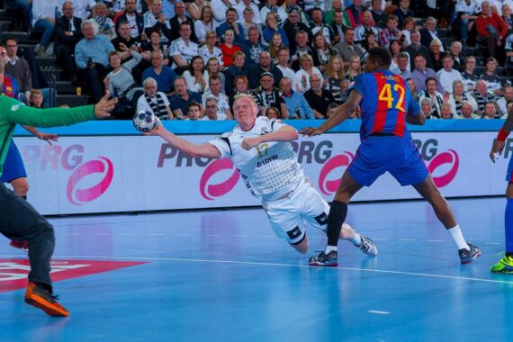 Ein Handballspieler in einem weißen Trikot springt und schießt den Ball in die Luft, während gegnerische Spieler in blauen und roten Trikots und ein grün gekleideter Torwart das Tor verteidigen, während die Zuschauer auf der Tribüne zusehen.