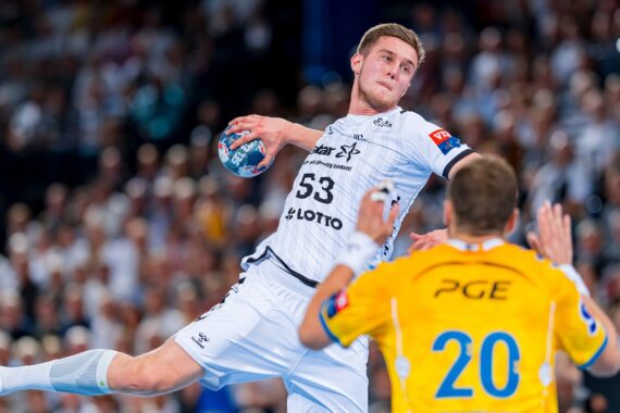 Ein Handballspieler in einem weißen Trikot bereitet sich in der Luft auf den Wurf vor, während ein Verteidiger in einem gelben Trikot mit der Nummer 20 die Arme hebt. Im Hintergrund ist eine unscharfe Menschenmenge zu sehen.