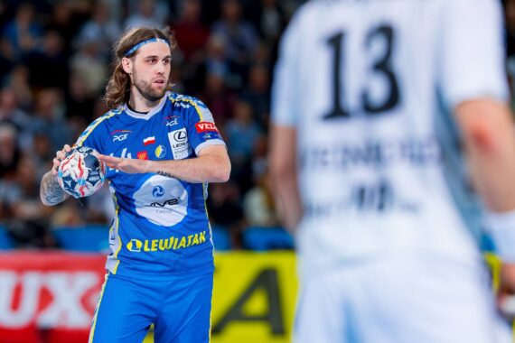 Ein Handballspieler in einem blau-gelben Trikot bereitet sich auf einen Spielzug vor, während er den Ball hält. Im Vordergrund ist ein verschwommener Spieler in einem weißen Trikot mit der Nummer 13 zu sehen. Die Szene spielt sich in einer belebten Halle ab.