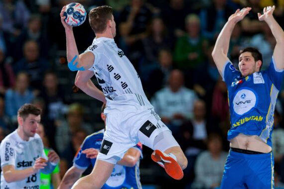 Ein Handballspieler in einem weißen Trikot springt, um den Ball zu werfen, während ein Verteidiger in einem blauen Trikot die Arme zum Blocken hebt.