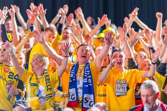 Eine große Menge begeisterter Sportfans in gelben Hemden und Schals jubelt mit erhobenen Armen und zeigt damit Begeisterung und Einigkeit bei einer Veranstaltung.