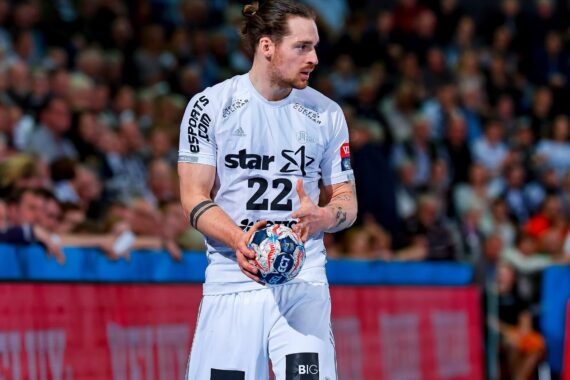 Ein männlicher Handballspieler, der ein weißes Trikot mit der Nummer 22 trägt, hält während eines Spiels einen bunten Handball, mit einer unscharfen Menschenmenge im Hintergrund.