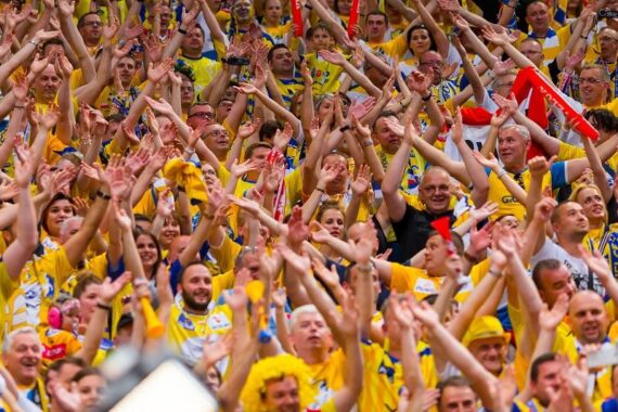 Eine große Menge begeisterter Fans in gelben und blauen Hemden jubelt mit erhobenen Händen bei einem Sportereignis und zeigt damit Begeisterung und Einigkeit. Einige Leute tragen Hüte und Perücken, und eine rot-weiße Flagge ist zu sehen.