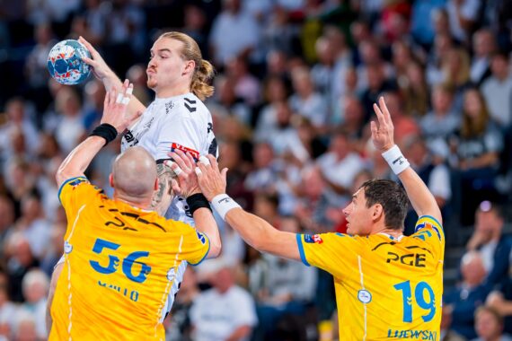 Ein Handballspieler in einem weißen Trikot springt und bereitet sich auf den Wurf vor, während zwei Gegner in gelben Trikots ihn mit erhobenen Armen verteidigen. Der Hintergrund zeigt eine Zuschauermenge in einem Stadion.