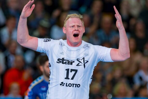 Ein Handballspieler, der ein weißes Trikot mit der Nummer 17 trägt, hebt während eines Spiels beide Arme und schreit, während im Hintergrund eine unscharfe Menschenmenge und ein anderer Spieler zu sehen sind.