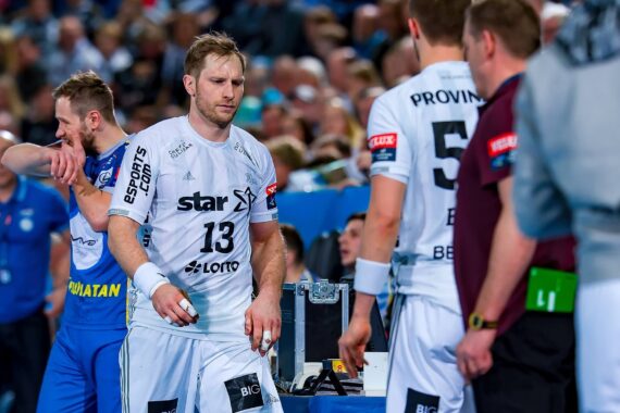 Ein männlicher Handballspieler in einem weißen Trikot mit der Nummer 13 geht enttäuscht vom Spielfeld, während Mannschaftskameraden und Offizielle während eines Spiels in der Nähe stehen. Unscharfe Menschenmenge im Hintergrund.