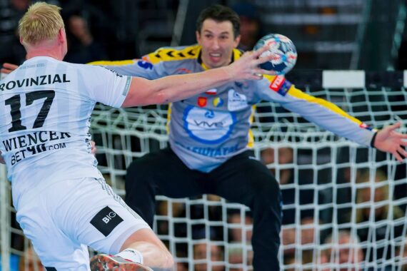 Ein Handballspieler in einem weißen Trikot springt und schießt auf ein Tor, während ein Torwart in Grau und Gelb seine Arme und Beine weit ausbreitet, um den Schuss zu blockieren. Die Aktion findet vor einem Tornetz statt.