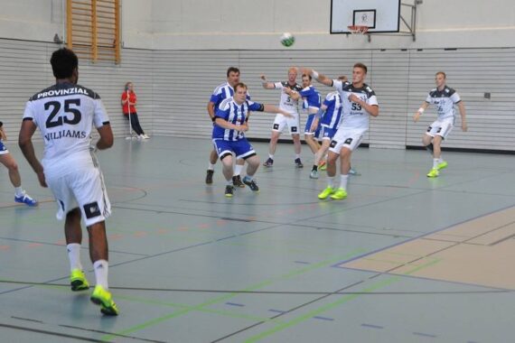 Eine Gruppe von Männern spielt ein Hallenhandballspiel. Spieler in weißen Trikots stehen Gegnern in blauen Trikots gegenüber, während sich ein Mitglied der blauen Mannschaft auf den Wurf vorbereitet, während die anderen zusehen und verteidigen. Das Spiel findet in einer Sporthalle statt.