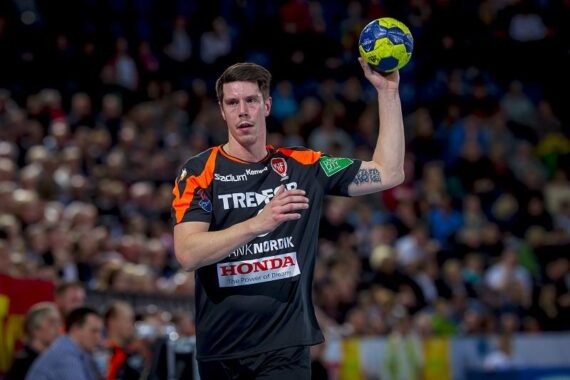 Ein männlicher Handballspieler in einem schwarz-orangefarbenen Trikot bereitet sich darauf vor, während eines Spiels einen blau-gelben Ball zu werfen, mit einer unscharfen Menschenmenge im Hintergrund.