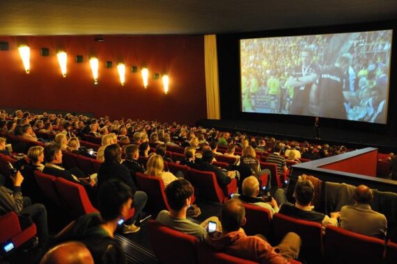 Eine große Gruppe von Menschen sitzt in einem Kino und verfolgt ein Sportereignis, das auf eine große Leinwand projiziert wird. Die Zuschauer sind auf die Leinwand konzentriert, und einige halten ihre Handys in der Hand.