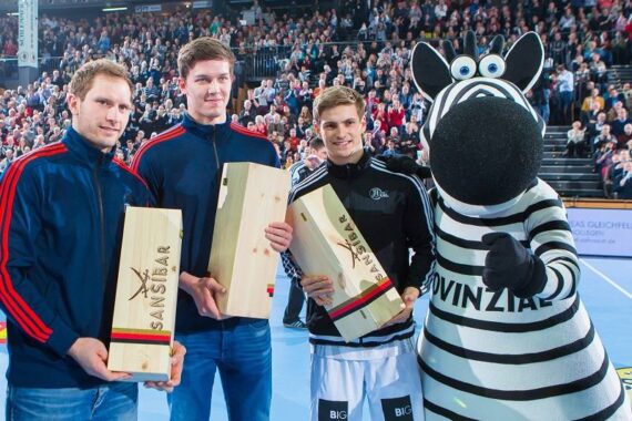Drei junge Männer mit hölzernen Trophäenkästen stehen neben einer Person in einem Zebra-Maskottchen-Kostüm auf einem Sportplatz, mit einer großen jubelnden Menge im Hintergrund.