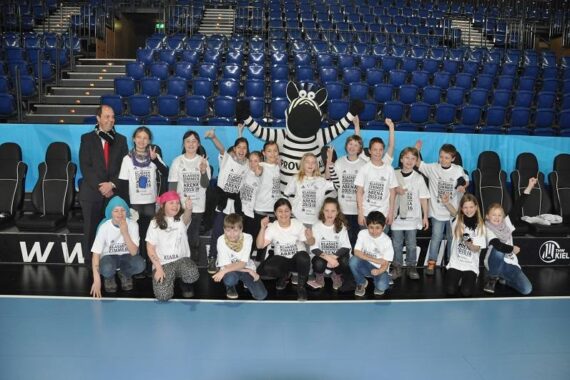 Eine Gruppe von Kindern und ein Erwachsener posieren zusammen in einer Arena und tragen passende weiße Hemden. Ein Zebra-Maskottchen steht hinter ihnen. Im Hintergrund sind blaue Sitzreihen zu sehen, und alle wirken fröhlich und aufgeregt.