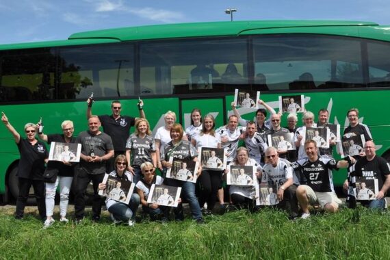 Eine Gruppe von Menschen posiert vor einem grünen Bus, hält Zeitungen oder Zeitschriften hoch und lächelt. Sie stehen im Gras und scheinen zu feiern oder aufgeregt zu sein, wobei viele einen Finger heben oder winken.
