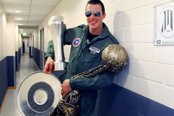 Ein Mann in einem grünen Fliegeranzug und mit Sonnenbrille steht in einem Flur, lächelt und hält drei große Sporttrophäen in der Hand, darunter eine runde, silberne und zwei hohe, metallene. An der Wand ist ein Schild mit der Aufschrift U3 zu sehen.