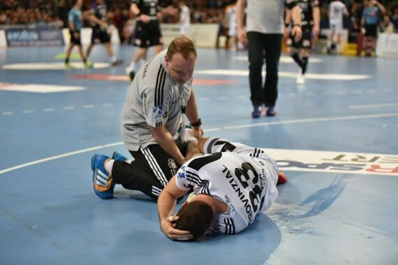 Ein Sportmediziner kümmert sich um einen verletzten Handballspieler, der auf dem Spielfeld liegt und sich den Kopf vor Schmerzen hält, während im Hintergrund andere Spieler und Zuschauer zu sehen sind.