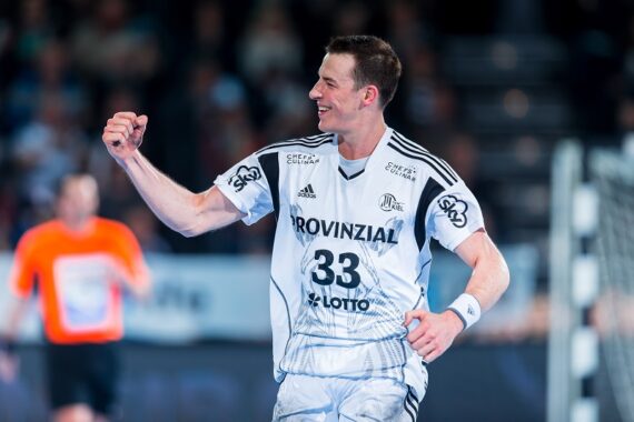 Ein männlicher Handballspieler, der ein weißes Trikot mit der Nummer 33 trägt, jubelt mit einem Fistpump. Er lächelt, und im Hintergrund ist ein verschwommener Schiedsrichter in einem orangefarbenen Hemd zu sehen.