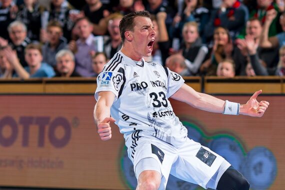 Ein männlicher Handballspieler in einem weißen Trikot feiert leidenschaftlich mit geballten Fäusten und offenem Mund während eines Spiels, während im Hintergrund eine jubelnde Menge zusieht.