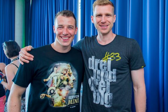 Zwei lächelnde Männer stehen nebeneinander vor einem blauen Vorhang. Der Mann auf der linken Seite trägt ein schwarzes T-Shirt mit einem grafischen Muster, der Mann auf der rechten Seite ein dunkelgraues T-Shirt mit gelber und weißer Schrift.