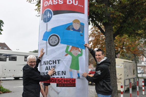 Zwei lächelnde Menschen stehen neben einem großen Straßenplakat, das eine Karikatur eines Mädchens zeigt, das ein blaues Auto mit einem überraschten Fahrer darin anhebt. Das Plakat hat einen fettgedruckten deutschen Text und ist an einem Mast in der Nähe einer Straße mit Bäumen und Fahrzeugen angebracht.