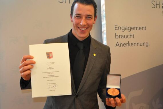 Ein Mann im Anzug lächelt, während er in der einen Hand eine Urkunde und in der anderen Hand eine Medaille in einem offenen Etui hält. Auf einem Schild im Hintergrund steht "Engagement braucht Anerkennung".