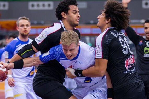 Drei männliche Handballspieler kämpfen während eines Spiels aggressiv um den Ball, wobei zwei Spieler in schwarzen Trikots einen Spieler in einem weiß-blauen Trikot blockieren. Die Szene fängt intensiven Körperkontakt und Konzentration ein.