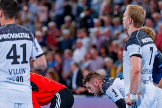 Drei männliche Handballspieler in weißen Trikots mit der Aufschrift PROVINZIAL auf dem Rücken stehen auf einem Hallenplatz und wirken konzentriert und intensiv, während das Publikum im Hintergrund verschwommen ist.