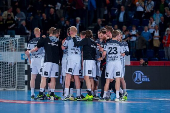 Eine Herren-Handballmannschaft in weiß-schwarzen Trikots kauert auf einem Hallenplatz zusammen, während im Hintergrund Zuschauer von der Tribüne aus zusehen.