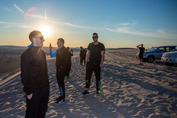 Mehrere Personen stehen bei Sonnenuntergang in einer Sandwüste, einige lächeln und unterhalten sich, während eine Person ein Foto mit dem Handy macht und eine andere in der Nähe geparkter SUVs in die Ferne schaut. Die Sonne steht tief am Horizont.