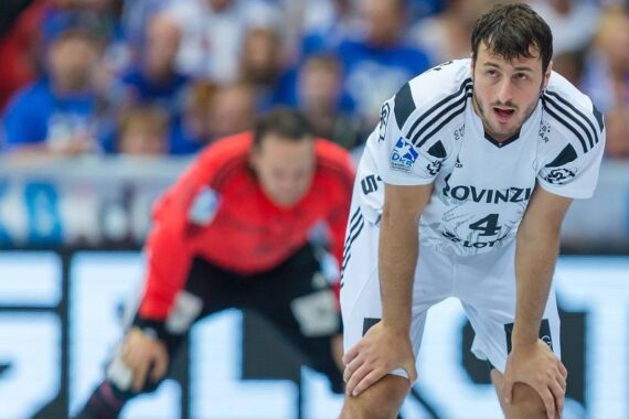 Ein männlicher Handballspieler in einem weißen Trikot mit der Aufschrift SLOVENIJA lehnt sich mit den Händen auf den Knien nach vorne, während ein Torwart in einem roten Trikot unscharf im Hintergrund steht. Hinter ihnen ist ein Publikum zu sehen.