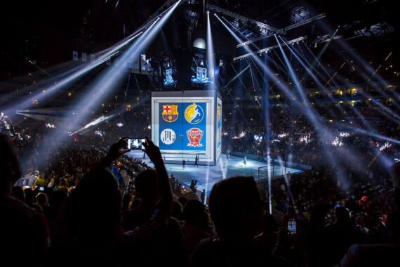 Eine große, mit Zuschauern gefüllte Halle mit einer hell erleuchteten Bühne in der Mitte, auf der die Logos von vier Sportmannschaften auf Bildschirmen zu sehen sind, umgeben von dramatischen weißen Lichtkegeln und Menschen, die Fotos machen.