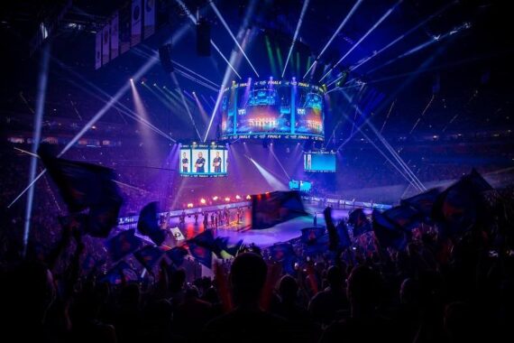 Eine große Menschenmenge schwenkt Fahnen in einer überdachten E-Sport-Arena mit bunten Lichtern und einem riesigen Bildschirm, der die Spielerprofile über der Bühne anzeigt. Die Atmosphäre ist lebhaft und energiegeladen.