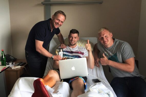 In einem Krankenhauszimmer lächeln drei Männer in die Kamera. Einer sitzt mit einem bandagierten Bein auf einem Krankenhausbett und benutzt einen Laptop. Die beiden anderen stehen und sitzen neben ihm, zeigen die Daumen nach oben und halten Geschenke.