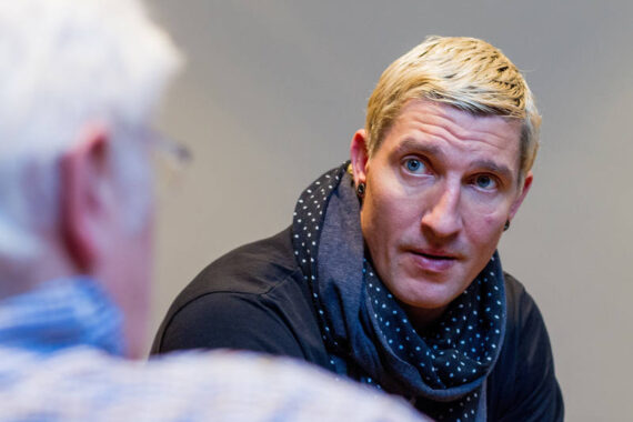 Eine Person mit kurzen blonden Haaren und einem dunklen Tupfenschal blickt aufmerksam auf eine andere Person im Vordergrund, die unscharf ist. Der Hintergrund ist schlicht und neutral.