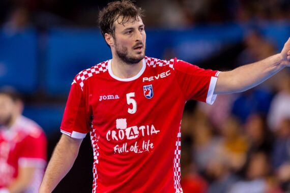 Ein männlicher Handballspieler in einem roten kroatischen Mannschaftstrikot mit der Nummer 5 zeigt während eines Spiels den Daumen nach oben. Der Hintergrund zeigt eine unscharfe Zuschauermenge und blaue Arenasitze.