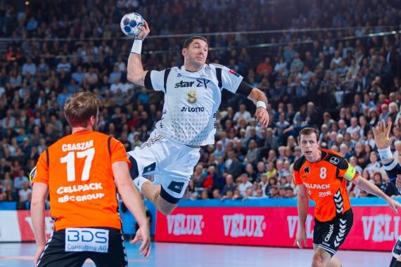 Ein Handballspieler in einem weißen Trikot springt in die Luft, um den Ball zu werfen, während zwei Spieler in orangefarbenen Trikots den Ball verteidigen. Eine große Menschenmenge verfolgt das intensive Spiel in der Arena.