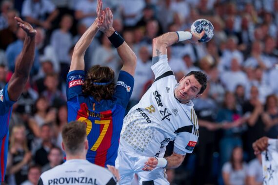 Ein Handballspieler in einem weißen Trikot springt und bereitet sich auf den Wurf vor, während ein Verteidiger in einem blau-roten Trikot versucht, ihn abzuwehren. Die Aktion findet in einer überfüllten Halle statt.