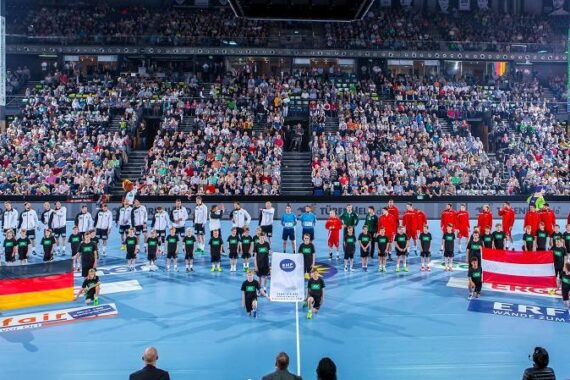 Zwei Handball-Nationalmannschaften stehen in Reihen auf einem blauen Spielfeld, wobei jedes Kind die Flagge seines Landes vor sich hält, während ein großes Publikum die Halle füllt.