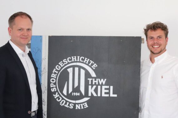 Zwei Männer in weißen Hemden stehen lächelnd zu beiden Seiten eines schwarzen Schildes mit dem Logo und dem Text THW Kiel 1904 Ein Stück Sportgeschichte vor einem schlichten weißen Hintergrund.