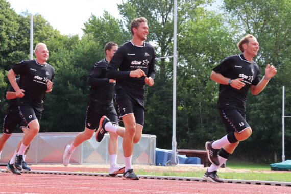 Vier Männer in schwarzen Sportuniformen joggen auf einer roten Laufbahn im Freien, umgeben von grünen Bäumen und blauem Himmel. Sie scheinen gemeinsam zu trainieren und lächeln, während sie laufen.