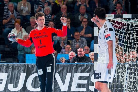 Ein Handballtorhüter in einem roten Trikot hebt jubelnd die Faust, während er einem gegnerischen Spieler in einem weißen Trikot in der Nähe des Tores gegenübersteht, während im Hintergrund eine Zuschauermenge zusieht.