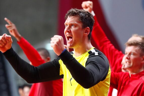 Ein männlicher Athlet im gelben Trikot ballt die Fäuste und jubelt, während seine Mannschaftskameraden in Rot im Hintergrund enthusiastisch jubeln.