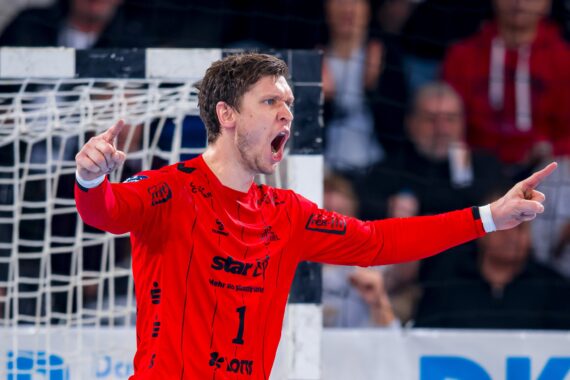 Ein männlicher Handballtorhüter in einem roten Trikot steht vor dem Torpfosten, ruft und zeigt mit beiden Händen während eines intensiven Moments in einem Spiel, mit Zuschauern im Hintergrund.