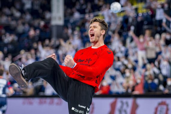 Ein männlicher Handballspieler in einem roten Trikot und einer schwarzen Hose feiert energisch auf dem Spielfeld, hebt die Faust und schreit, während im Hintergrund eine verschwommene Menge jubelt.